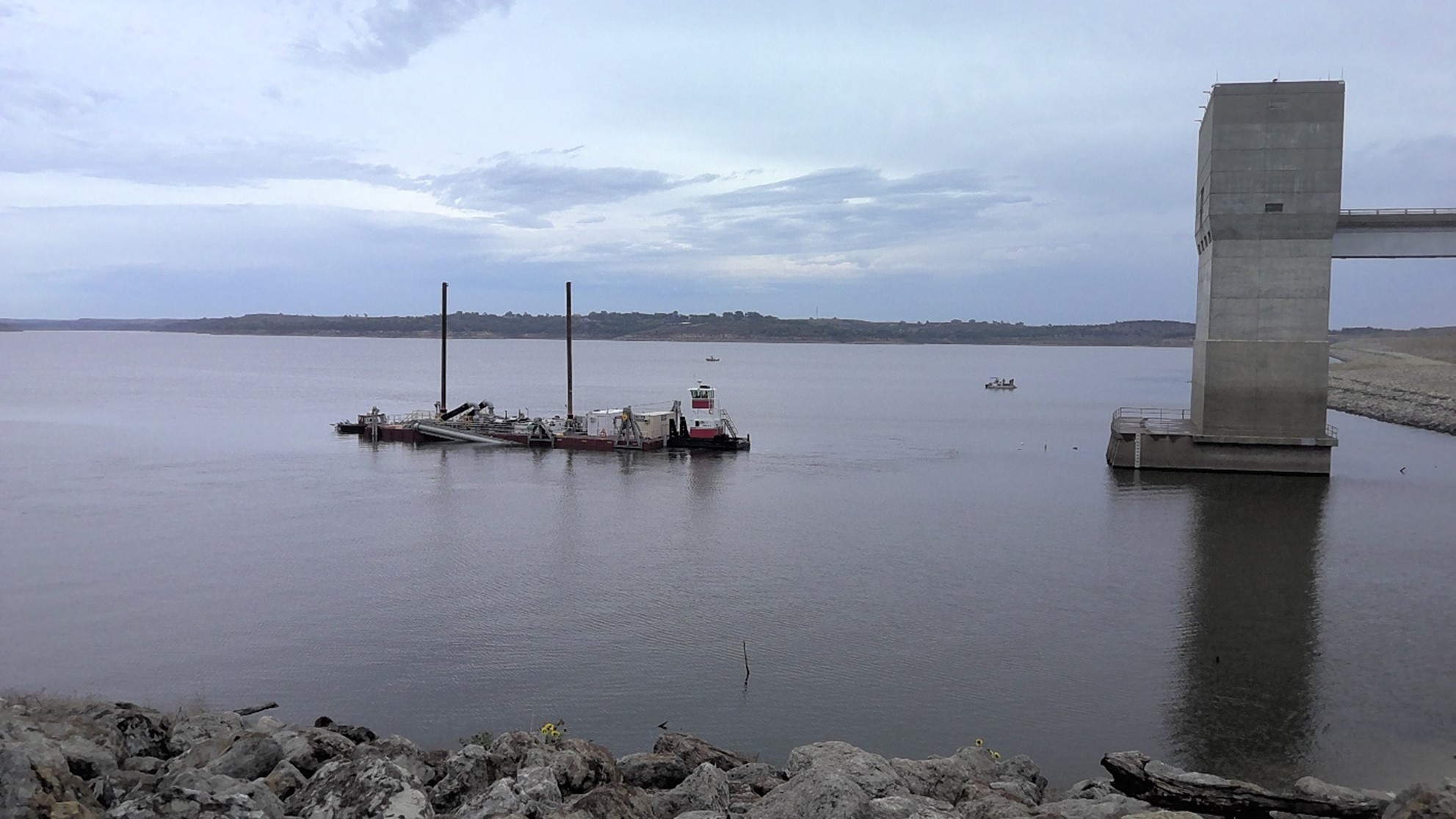 Large reservoir scene with a dredging barge positioned in open water. The barge includes tall vertical poles, equipment, and a small control cabin. A tall concrete intake structure stands on the right side of the image. Rocky shoreline appears in the foreground, with cloudy skies and distant hills in the background.