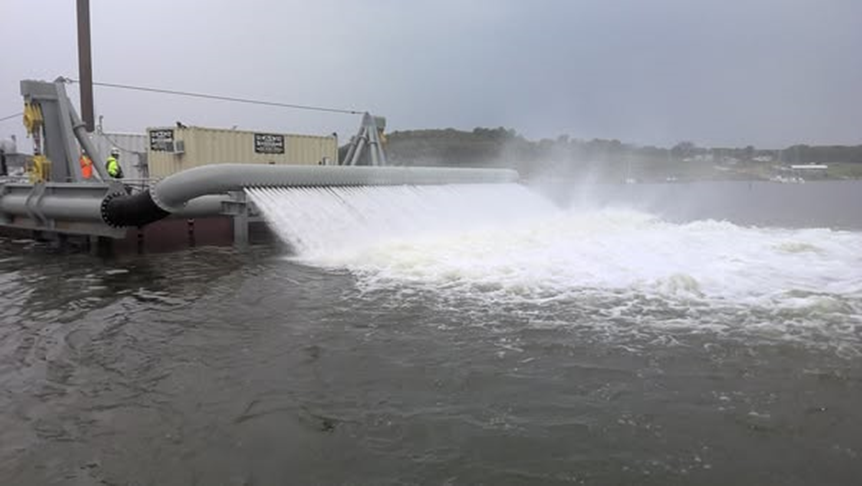 Close-up view of a sediment-release or water-discharge structure mounted on a barge. A curved pipe releases a high-velocity sheet of white, aerated water into the reservoir, creating turbulence. Workers in safety gear stand on the deck near equipment and containers.