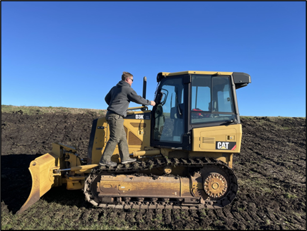 A person climbing into the cab of a yellow CAT D3K bulldozer parked on a dirt slope under a clear blue sky. Freshly graded soil surrounds the machine.