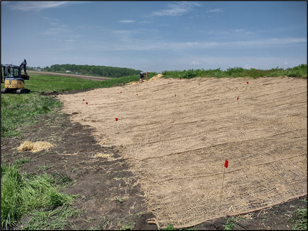 A newly constructed slope covered in erosion-control matting with small red flags marking plot locations. A compact excavator sits in the distance on the left, with grassy fields and trees surrounding the site under a blue sky.