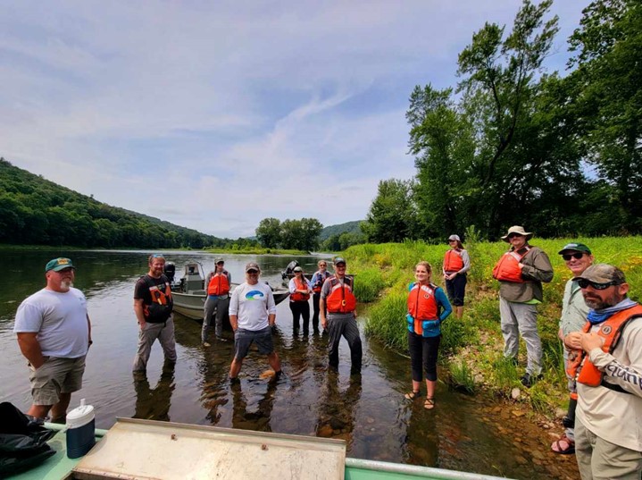 Group of people standing in shallow water along a riverbank, wearing life jackets and field gear. A boat is anchored nearby. Forested hills surround the river under a partly cloudy sky.