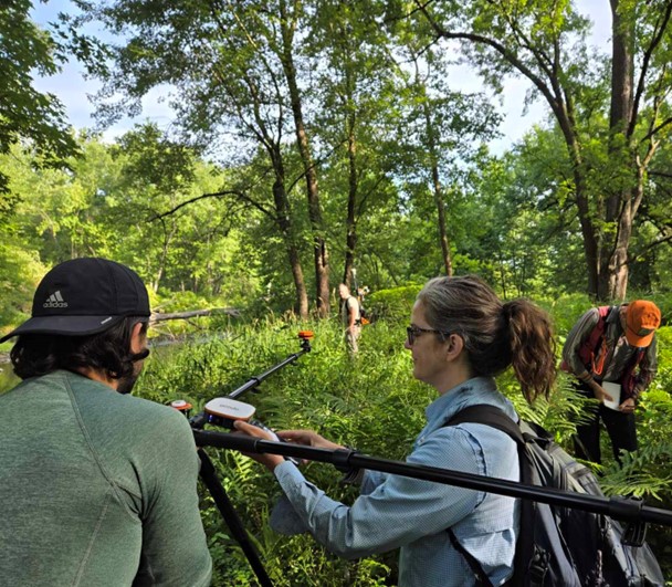Two field researchers using surveying poles and GPS equipment in a lush, green riverside forest. One person writes notes while another adjusts equipment. Dense vegetation and tall trees surround the team.