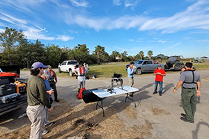 Logistics/safety meeting for UAS. At least eight people standing, surrounding a table with a drone on top of it. Pickup trucks are in the background.