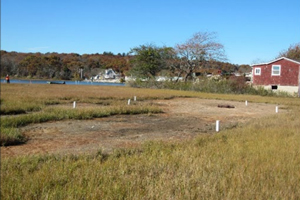 Photo of a grassy area with clearing outlines with periodic vertical white pipes, a red house and a lake in the background on a cloudless day.